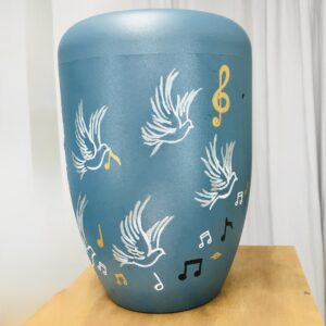 Die fertige türkise Workshop-Urne Taube mit goldenen, schwarzen und weißen Musiknoten neben weißen Tauben.