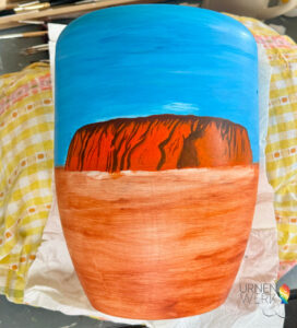 Urne mit einer Landschaftsmalerei, die einen orangen Felsen unter einem blauen Himmel zeigt, umgeben von einem braunen Boden.